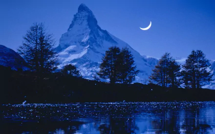 A serene evening in Switzerland features the Matterhorn illuminated by a crescent moon, surrounded by tranquil blue skies, trees, and reflections on a calm lake.