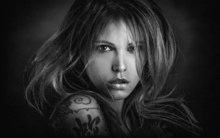 Black-and-white close-up of a tattooed female model with windblown hair and an intense gaze — HD PC desktop wallpaper background.
