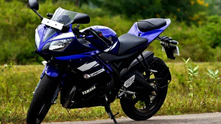 A striking Yamaha YZF-R15 motorcycle in vibrant blue, parked on a scenic roadside with greenery in the background, making for an impressive HD desktop wallpaper.