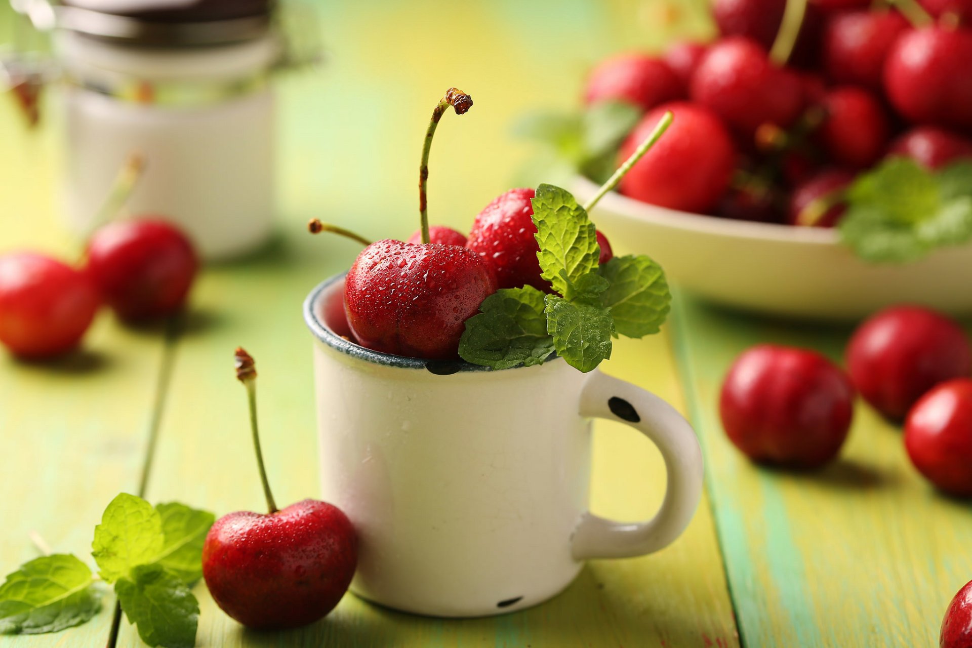 Close-up of fresh cherries and mint leaves arranged in a white mug on a colorful wooden surface, captured as a vibrant HD PC desktop wallpaper and background.