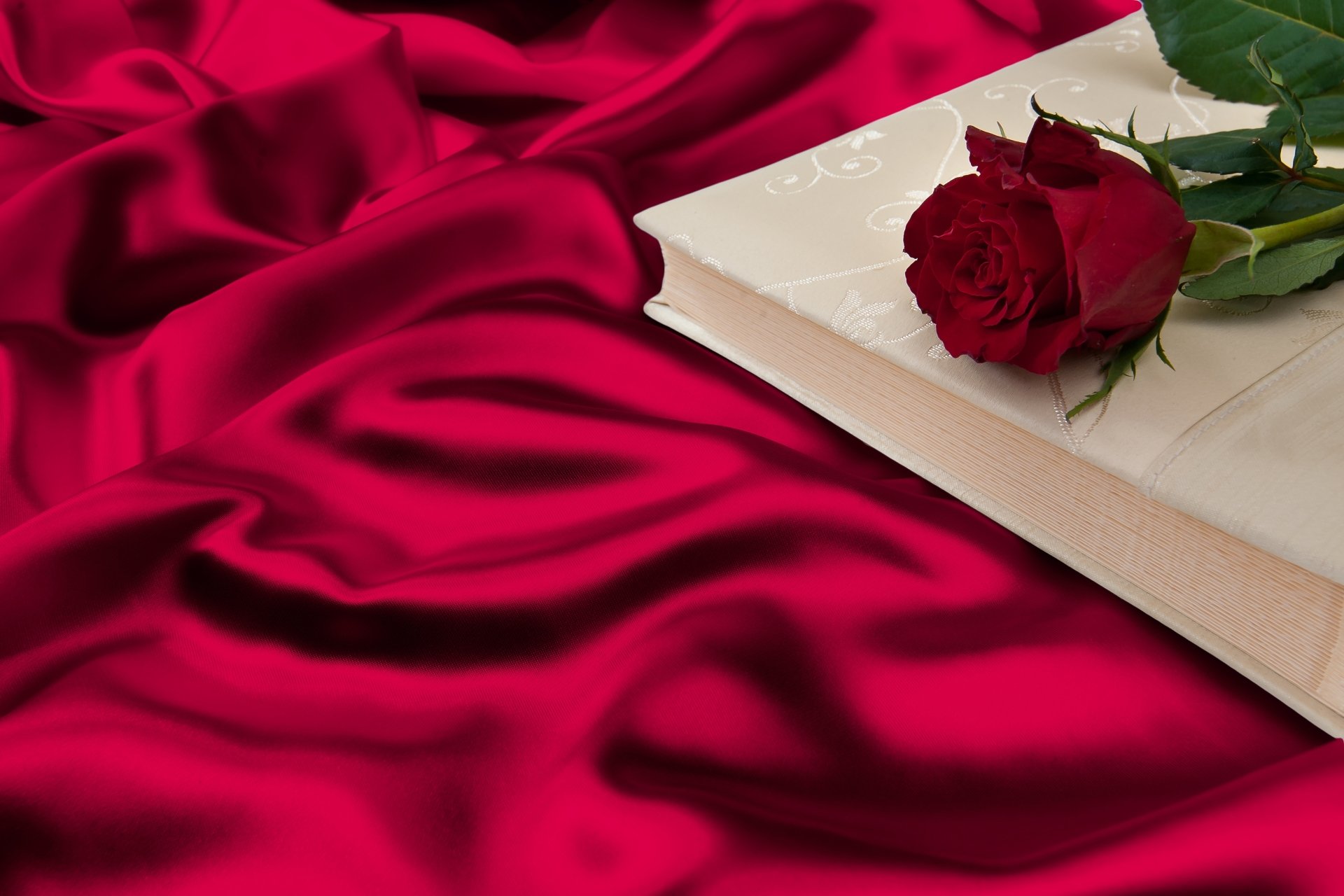 A vibrant red rose rests on a closed book atop rich red silk fabric, captured in a high-resolution 4K Ultra HD still life photography.