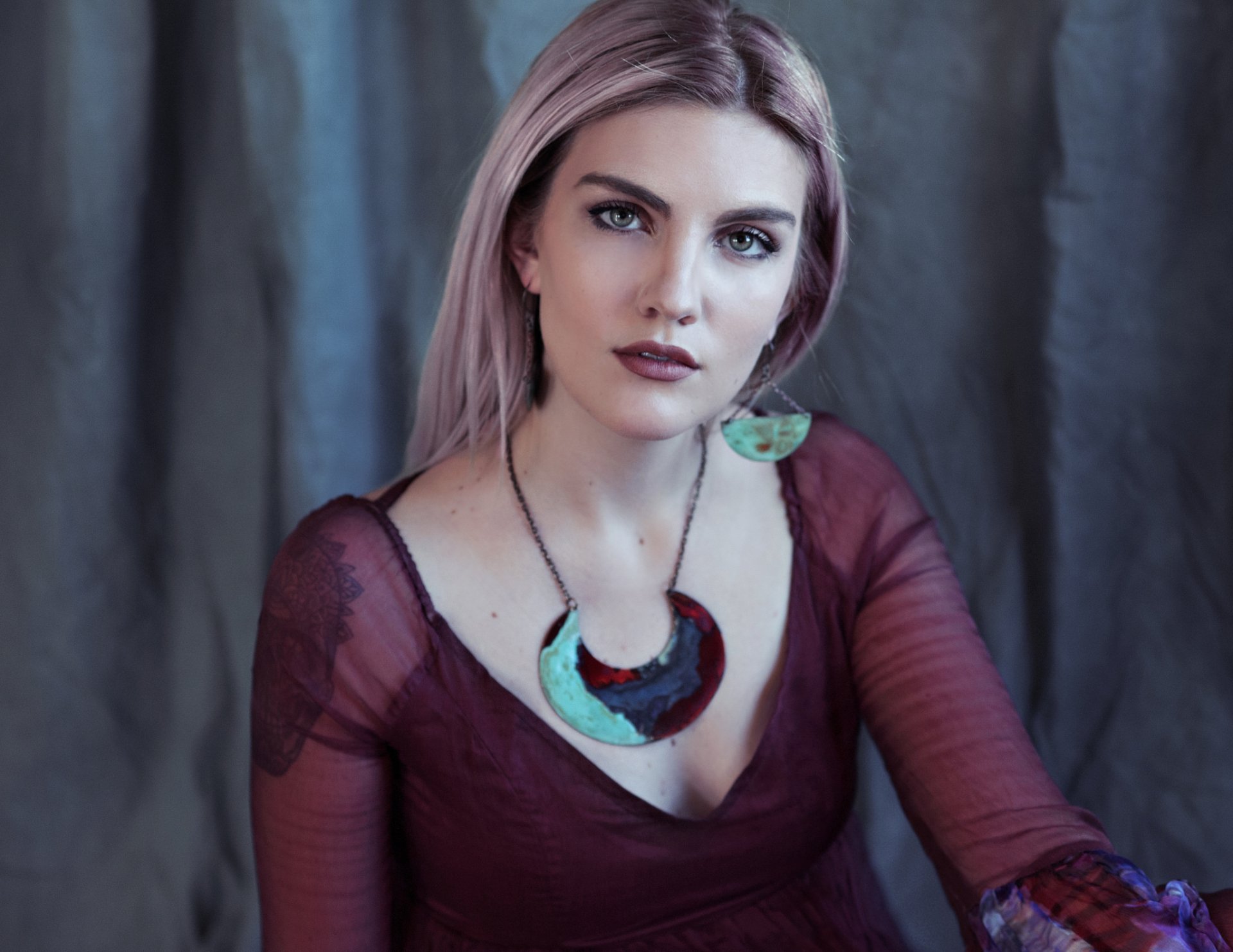 Blonde model with green eyes wearing turquoise earrings and a bold necklace, small chest tattoo visible, posed against a muted backdrop — HD PC desktop wallpaper/background