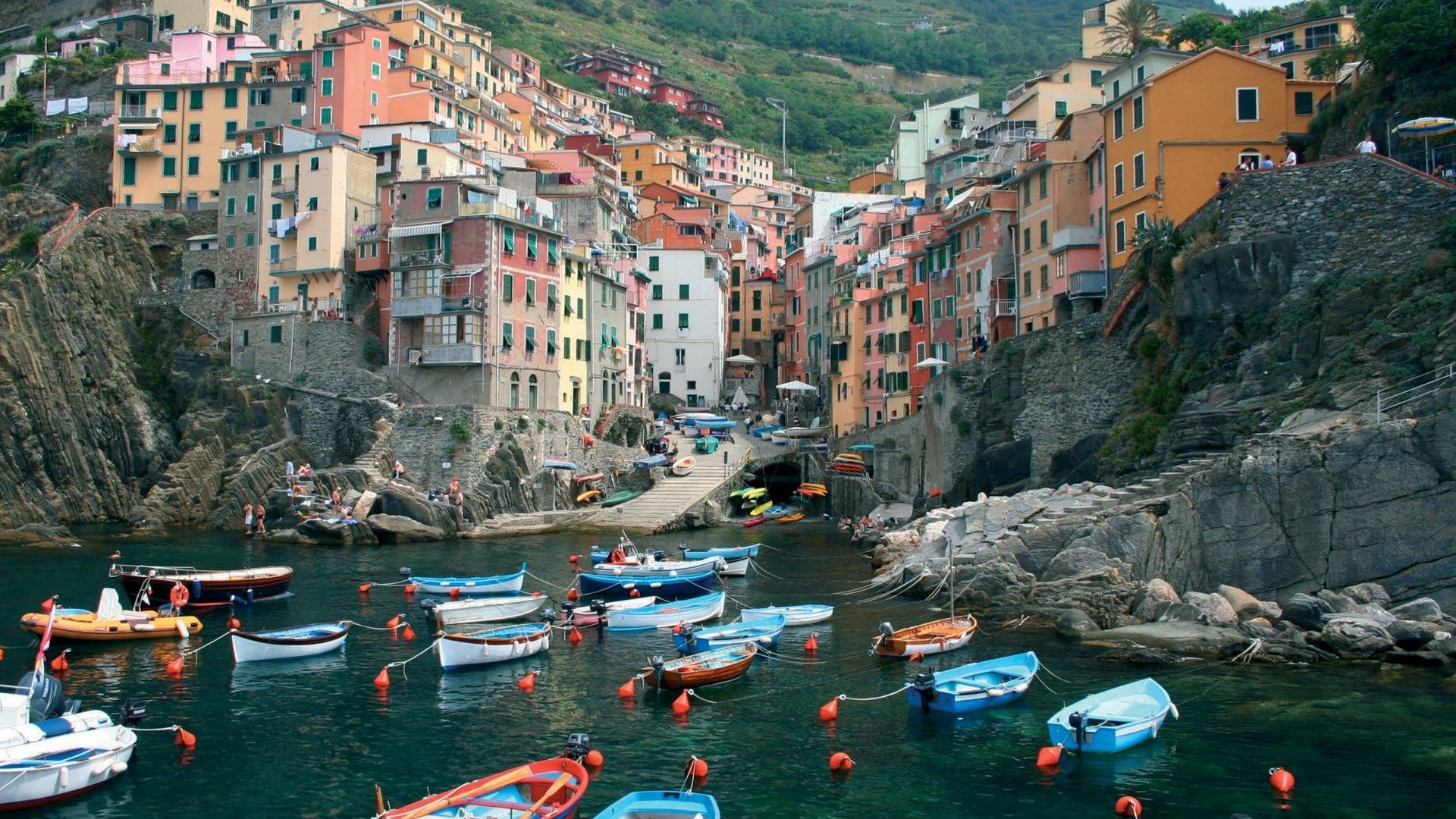 Colorful houses line the coast of Riomaggiore, Italy, with boats gently floating in the clear water, capturing the beauty of the Cinque Terre landscape.