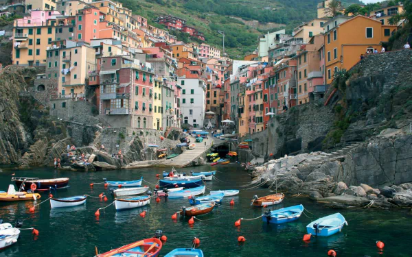 Colorful houses line the coast of Riomaggiore, Italy, with boats gently floating in the clear water, capturing the beauty of the Cinque Terre landscape.