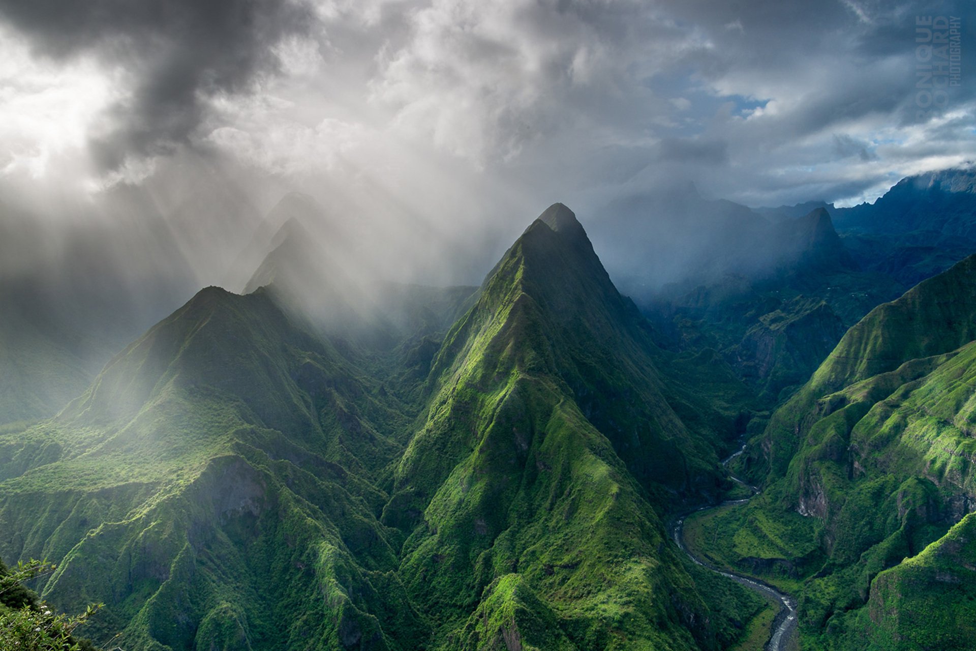 A breathtaking view of towering green mountains is illuminated by dramatic sunbeams breaking through foggy clouds, showcasing the beauty of nature in this HD wallpaper.