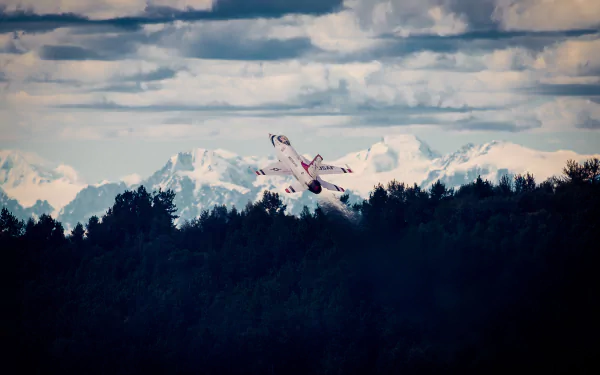 General Dynamics F-16 Fighting Falcon jet fighter from the U.S. Air Force Thunderbirds soars above a forest with snow-capped mountains under a cloudy sky.