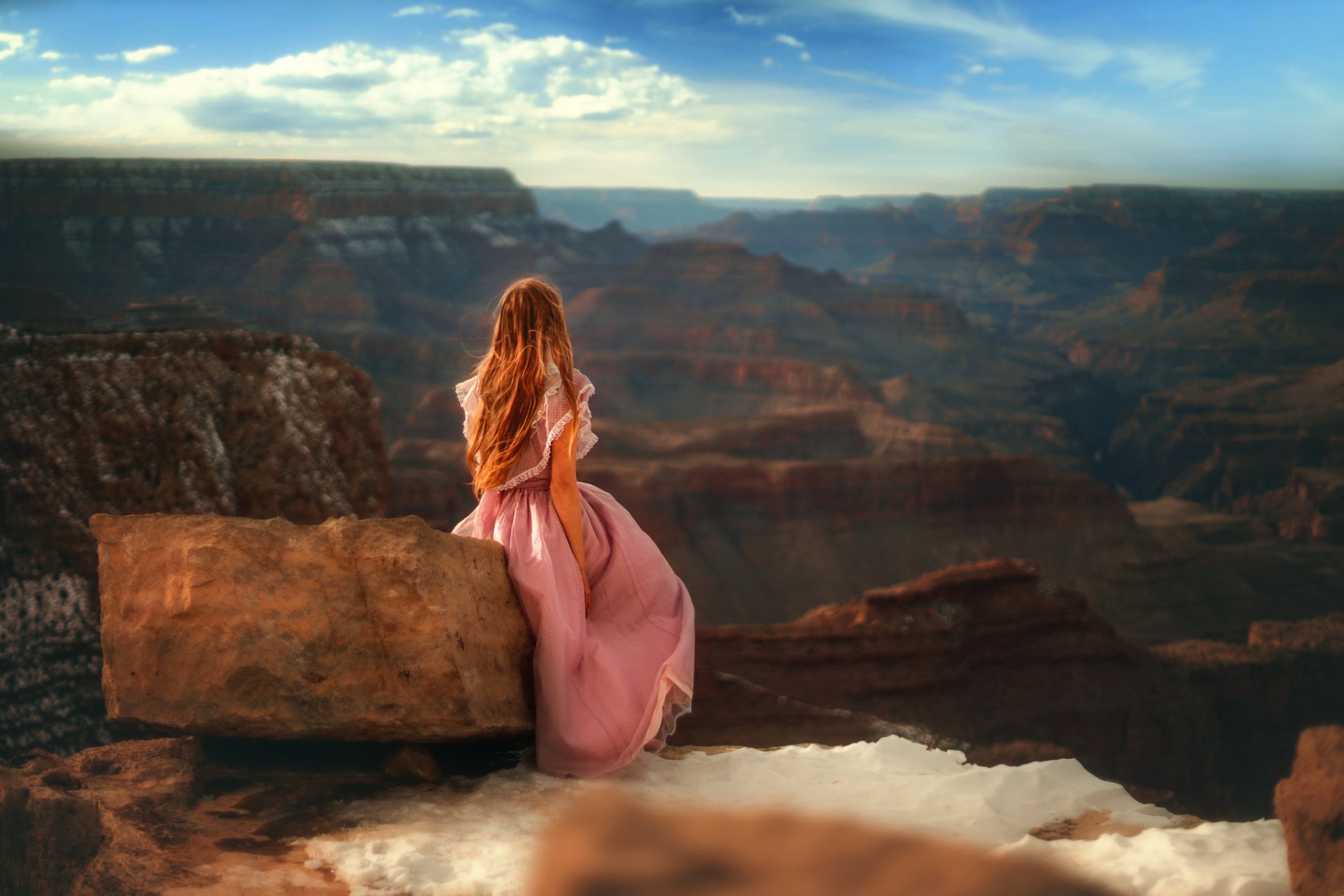 A woman with red hair in a pink dress sits on a rock, overlooking a vast canyon landscape. The scene, set outdoors under a blue sky, is captured in high-definition, making it a stunning desktop wallpaper.