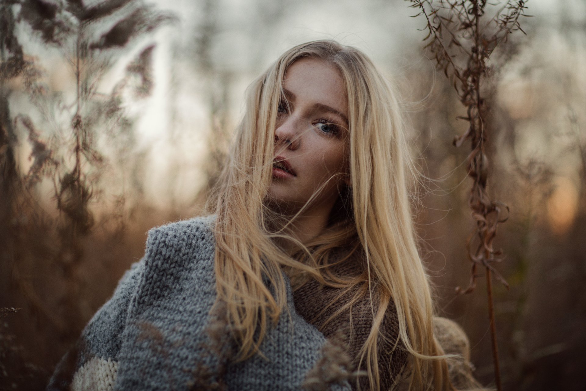 8K Ultra HD desktop wallpaper: close bokeh portrait of a blonde woman model with blue eyes wearing a cozy scarf, soft natural light and warm autumn tones.