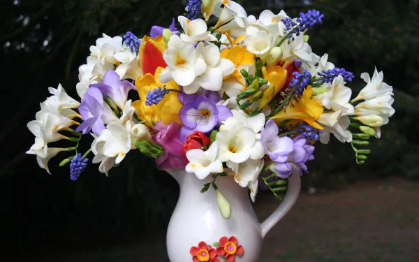 A vibrant bouquet of white freesias, crocuses, and other colorful flowers arranged in a decorative pitcher, set against a dark natural background.