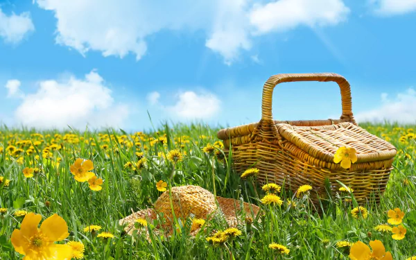 A picnic basket and straw hat rest in a vibrant yellow flower-filled field under a bright blue sky, capturing a cheerful spring scene.