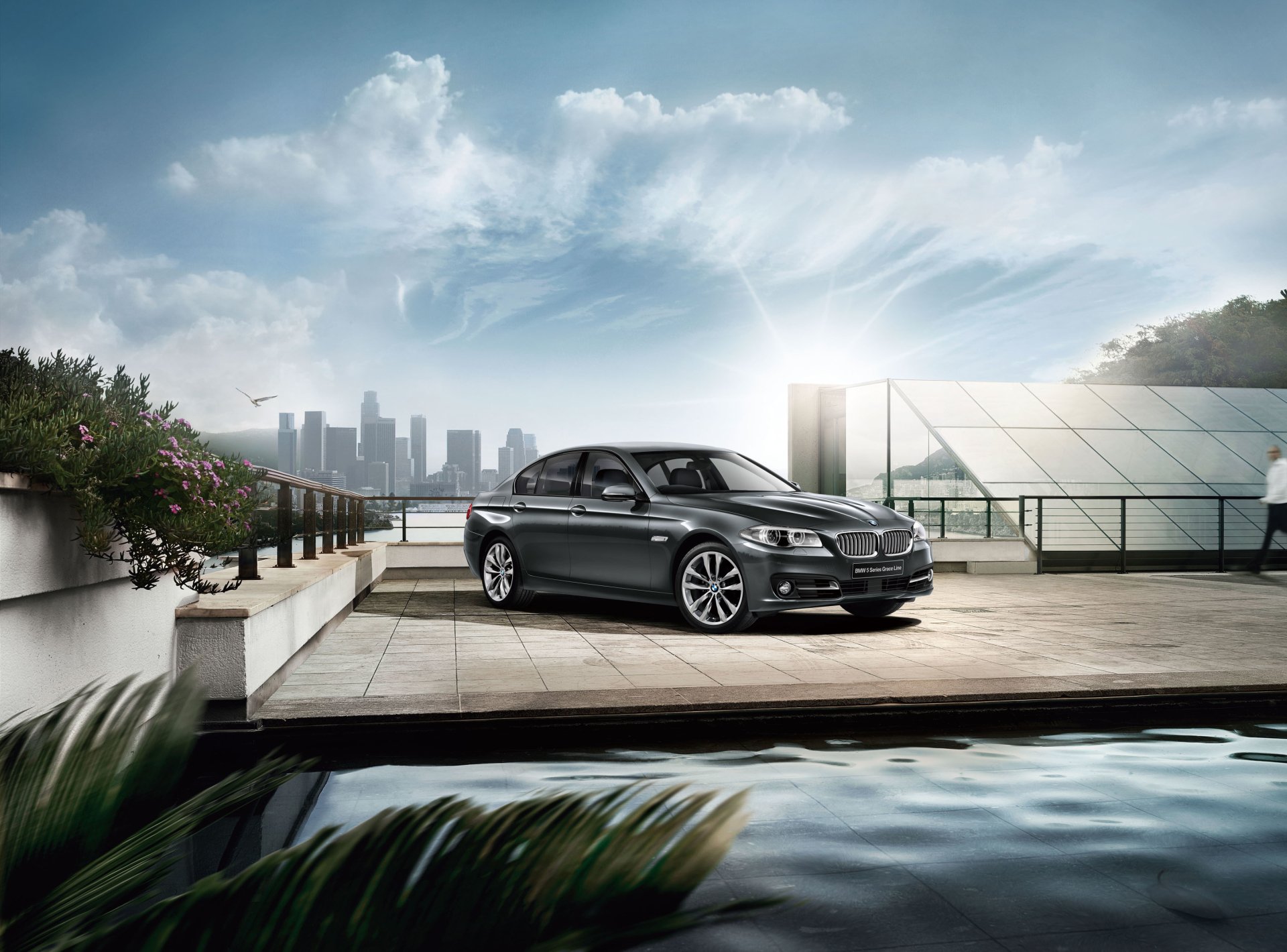 Black BMW 5 Series sedan parked on a waterfront terrace with city skyline and reflective pool — 2K Quad HD PC desktop wallpaper