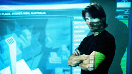Arms-crossed man with a bandana-like face covering standing before blue tactical displays — Mission: Impossible II movie HD PC desktop wallpaper/background.