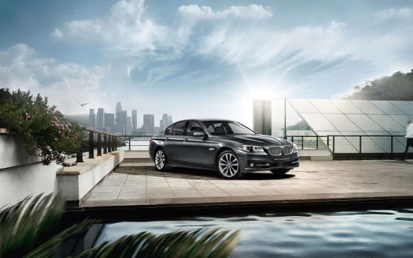 Black BMW 5 Series sedan parked on a waterfront terrace with city skyline and reflective pool — 2K Quad HD PC desktop wallpaper