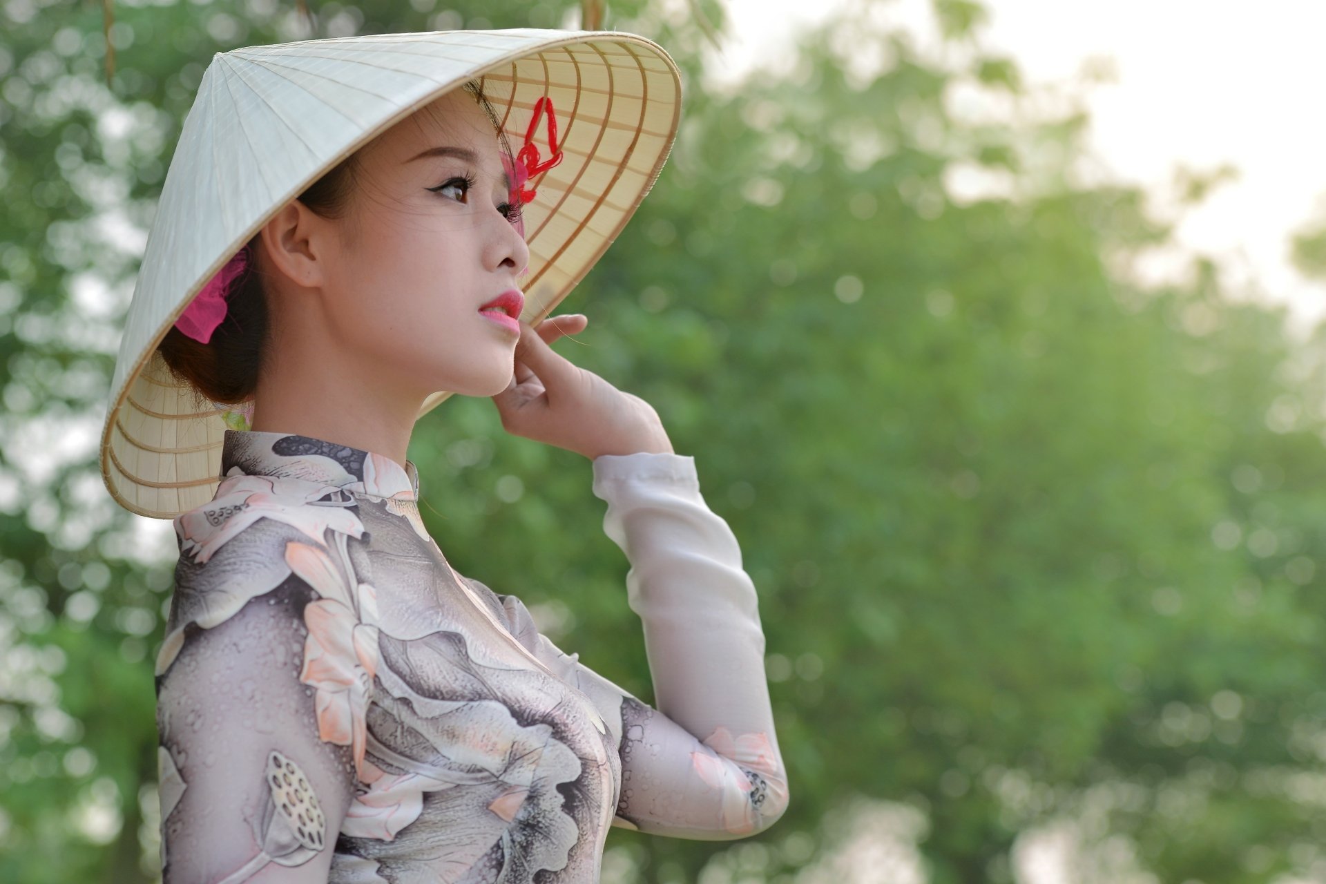 A brunette woman with brown eyes wears a traditional hat, posing gracefully against a soft bokeh background of greenery, creating a serene and captivating HD wallpaper.