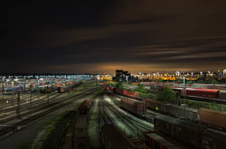 4K Ultra HD desktop wallpaper: nighttime railyard and train station with illuminated freight trains and wagons on multiple railroad tracks, glowing lights under a cloudy night sky.