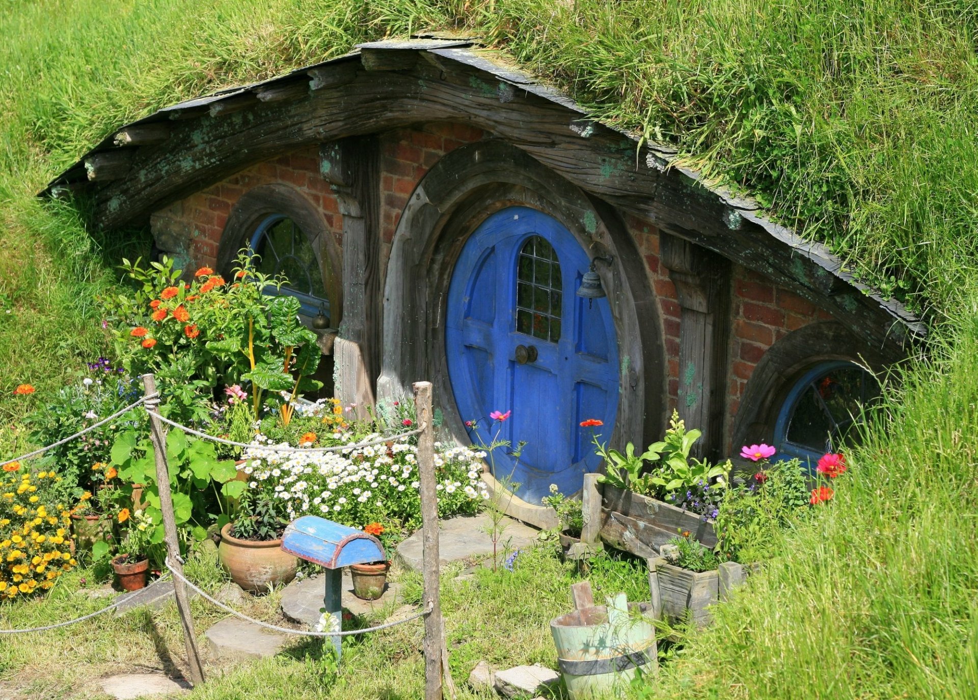 HD wallpaper of a charming man-made Hobbiton home with a round blue door, nestled in lush greenery and colorful flowers in the Comarca region.