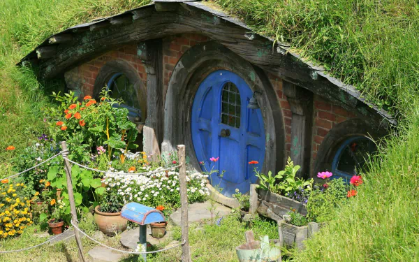 HD wallpaper of a charming man-made Hobbiton home with a round blue door, nestled in lush greenery and colorful flowers in the Comarca region.