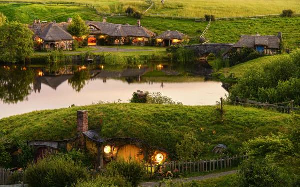Man-made Hobbiton village featuring charming, illuminated hobbit holes nestled in lush green hills by a reflective pond, captured in an HD PC desktop wallpaper.