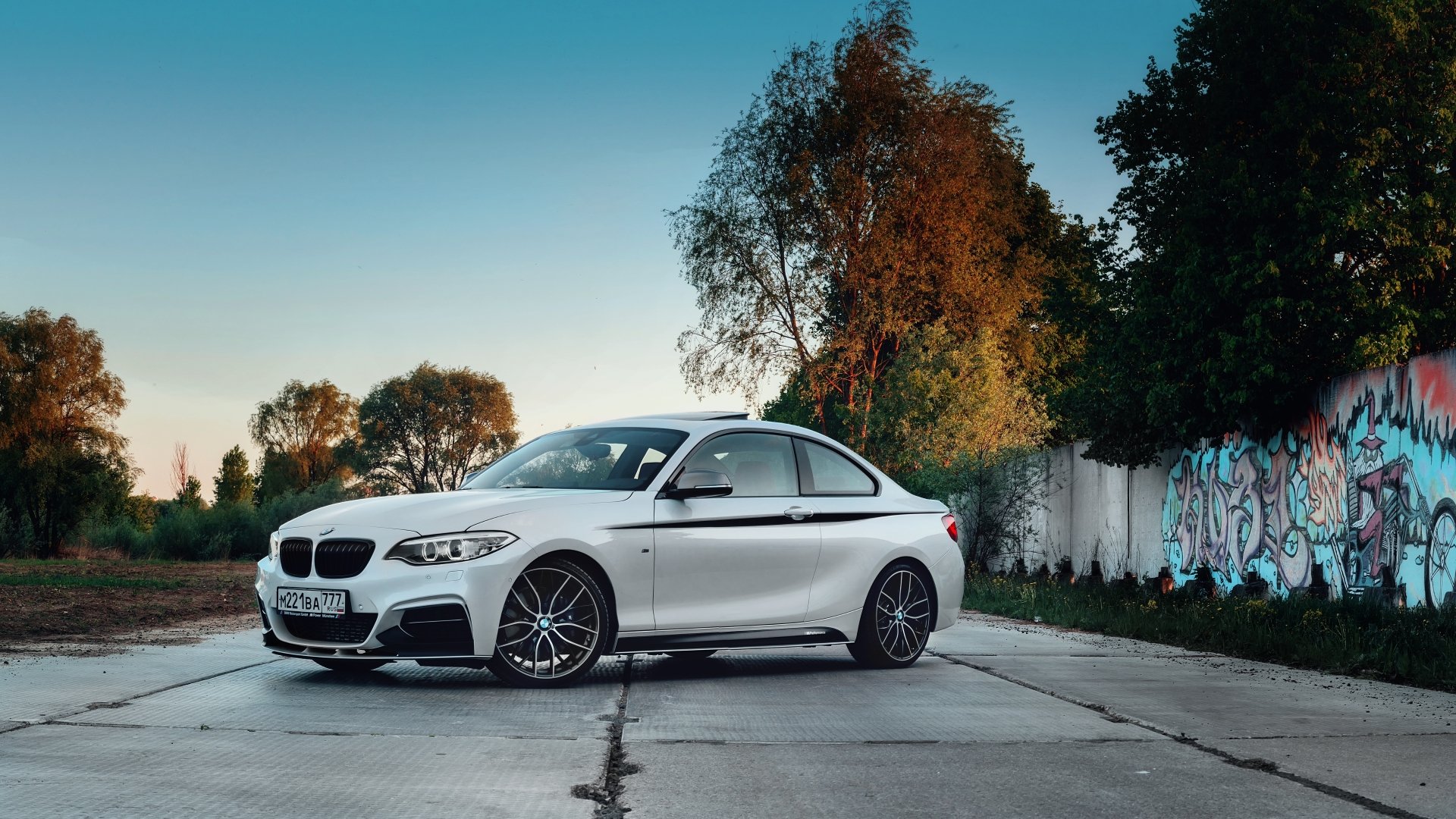 White BMW coupe parked on cracked concrete beside a graffiti wall at dusk — 5K Ultra HD PC desktop wallpaper and background showcasing a vehicle in an urban setting.