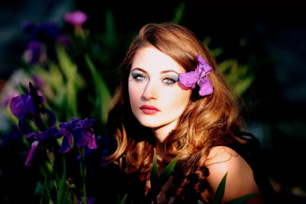 A redhead model with long hair, makeup, and lipstick poses surrounded by purple irises, with one flower tucked in her hair. The image is a high-definition desktop wallpaper.