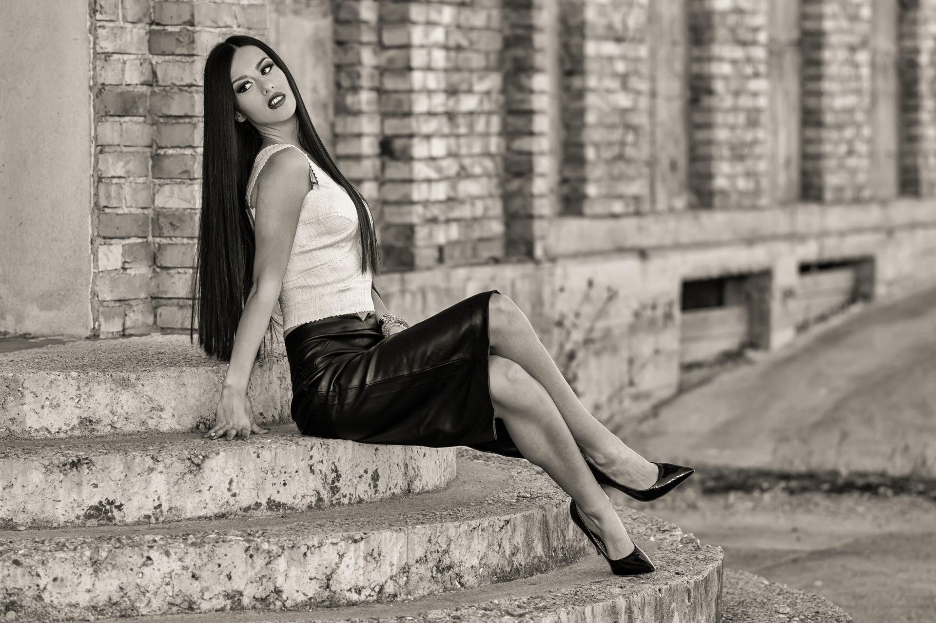 Black and white 4K Ultra HD wallpaper of a stylish woman with long hair and skirt, sitting on stairs against a brick wall, showcasing legs and confident pose.