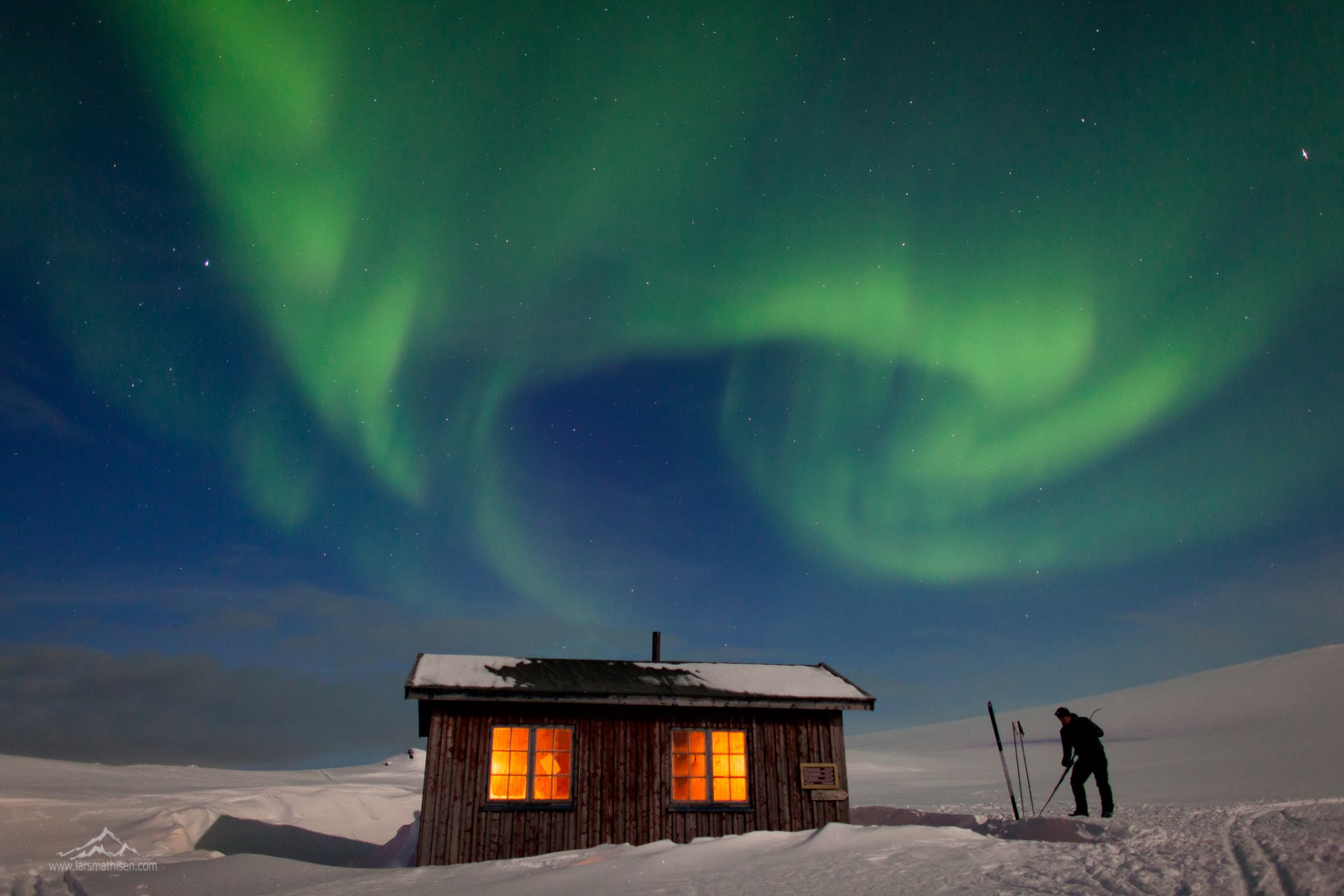 HD PC desktop wallpaper: snow-covered winter shed glowing with warm light beneath a vivid aurora borealis, a lone skier standing beside it.