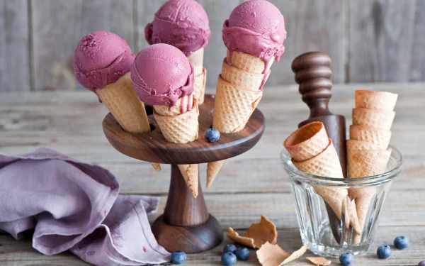 HD desktop wallpaper of a summer still life featuring scoops of pink ice cream in cones on a wooden stand, with additional cones in a glass jar and scattered blueberries.