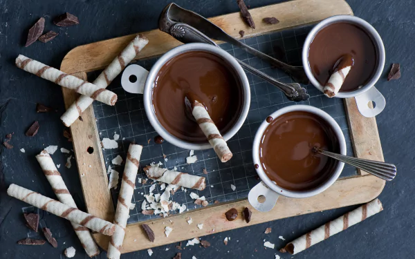 HD desktop wallpaper showcasing a still life of rich chocolate desserts, cups of melted chocolate, and striped wafer sticks arranged on a wooden board with scattered chocolate pieces.