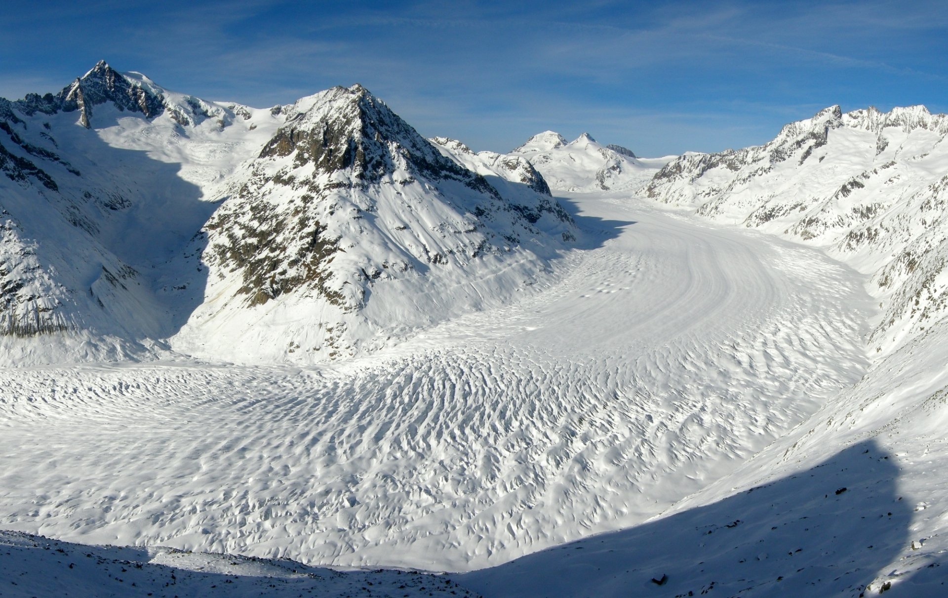 HD PC desktop wallpaper showing a sweeping glacier winding between snow-covered alpine peaks beneath a clear blue sky — nature background.