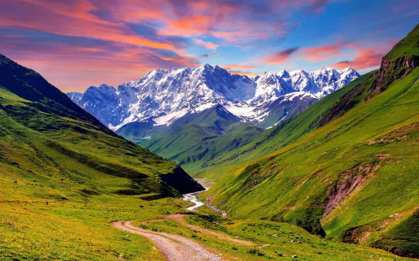 A breathtaking HD desktop wallpaper featuring a serene valley with a winding path and river, framed by lush green hills and majestic snow-capped mountains under a vibrant sunset sky.