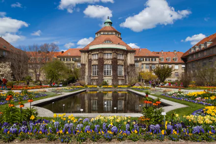 HD desktop wallpaper of a man-made garden featuring a grand historic building, colorful flower beds, a reflecting pond, and a vibrant blue sky with fluffy white clouds.