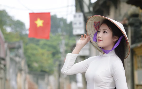 Vietnamese woman wearing a traditional Ao Dai and Asian conical hat stands in a street with a blurred Vietnamese flag in the background, captured in 4K Ultra HD.
