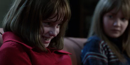 A still from The Conjuring 2 featuring Sterling Jerins as a young girl in a red shirt, smiling mischievously, with another girl in the background, creating an eerie atmosphere. HD desktop wallpaper.