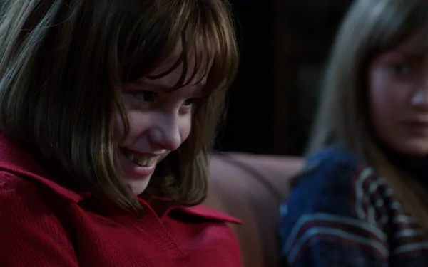 A still from The Conjuring 2 featuring Sterling Jerins as a young girl in a red shirt, smiling mischievously, with another girl in the background, creating an eerie atmosphere. HD desktop wallpaper.