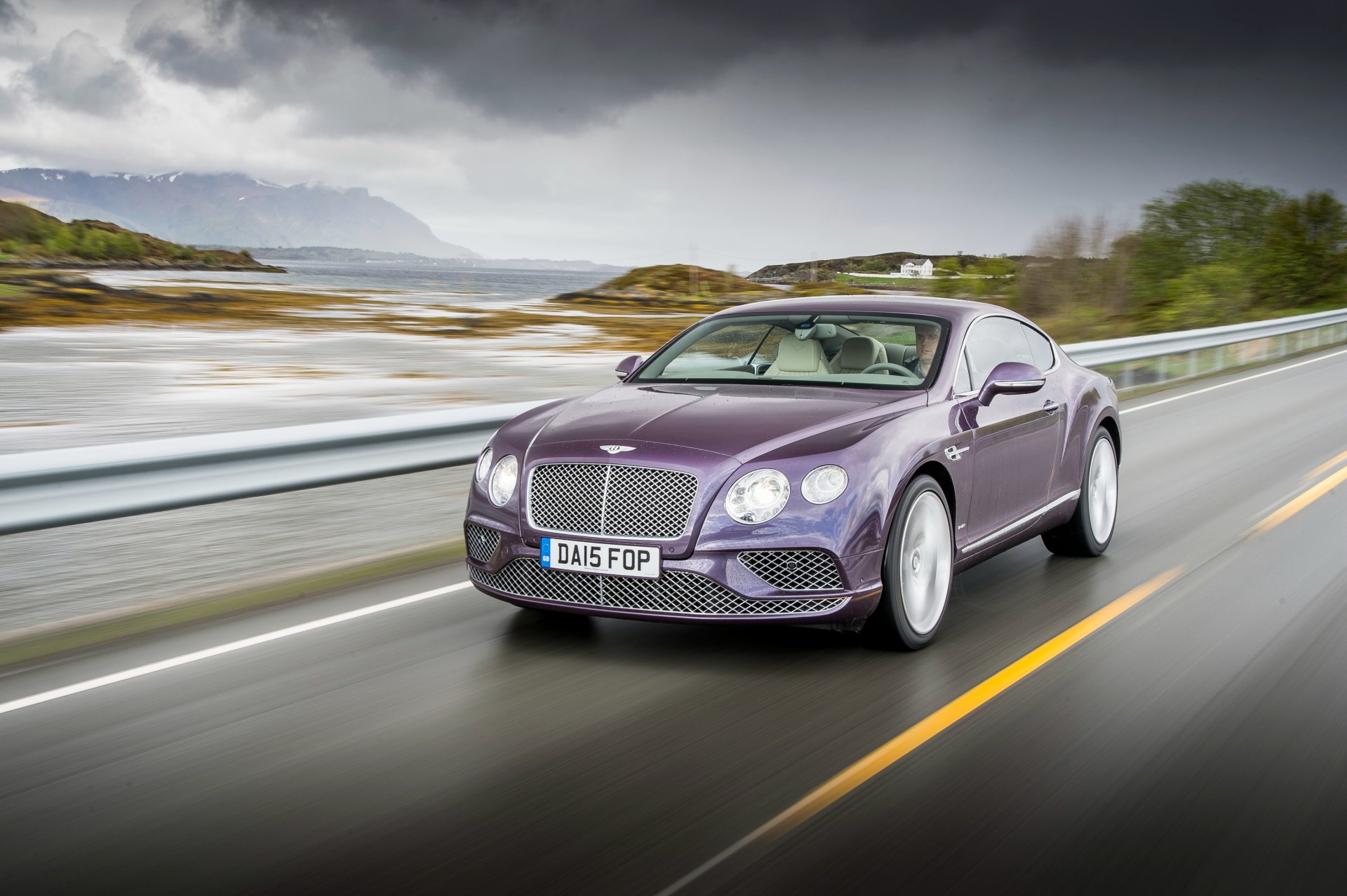 A purple Bentley Continental GT grand tourer driving on a coastal road with a cloudy sky, captured in 4K Ultra HD for a desktop wallpaper background.