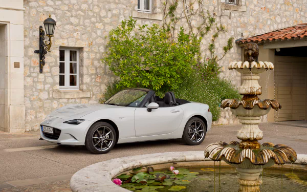 A sleek white Mazda MX-5 parked near a stylish stone building and a charming fountain, surrounded by lush greenery, captured in stunning 4K Ultra HD.