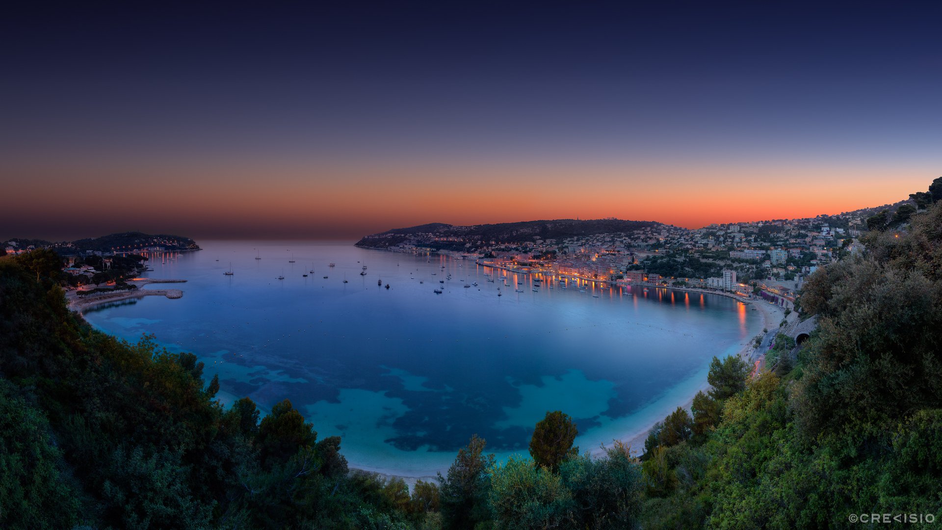 Night view of the man-made coastal town of Villefranche-sur-Mer, France, with city lights reflecting on calm waters under a clear twilight sky.