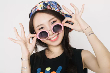 HD desktop wallpaper of an Asian brunette woman model with brown eyes, wearing braided hair, a colorful hat, pink sunglasses, and smiling playfully.