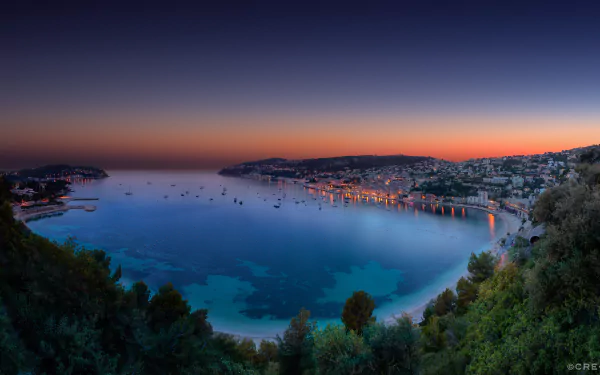 Night view of the man-made coastal town of Villefranche-sur-Mer, France, with city lights reflecting on calm waters under a clear twilight sky.