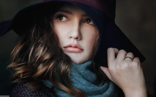 A close-up of a brunette woman with brown eyes, wearing a stylish scarf and a wide-brimmed hat, exuding elegance and poise against a soft backdrop.