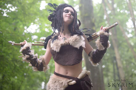 HD PC desktop wallpaper featuring a woman in detailed The Elder Scrolls V: Skyrim cosplay with fur and wooden props, set against a lush forest background.