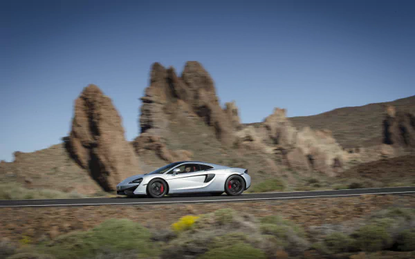 White McLaren 570GT supercar driving on a road with rocky mountains in the background, captured in a 4K Ultra HD PC desktop wallpaper and background.