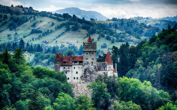HD wallpaper of Bran Castle, a man-made fortress nestled among lush green hills with misty mountains in the background.