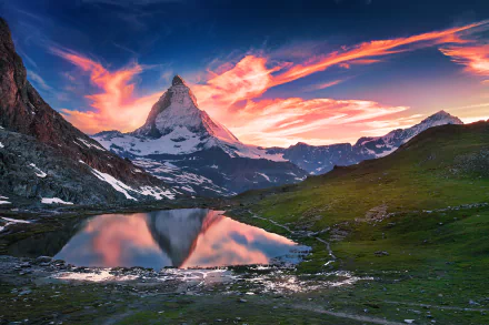 Dawn over the Matterhorn in Switzerland, with vibrant clouds reflected in a calm lake surrounded by mountains and lush green terrain.