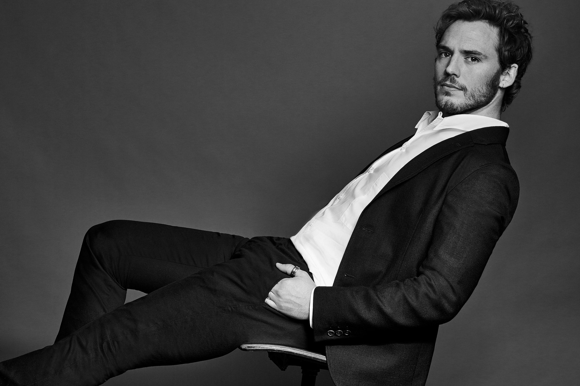 Black and white HD desktop wallpaper of English actor and celebrity Sam Claflin, dressed in a suit, reclining confidently against a dark backdrop.