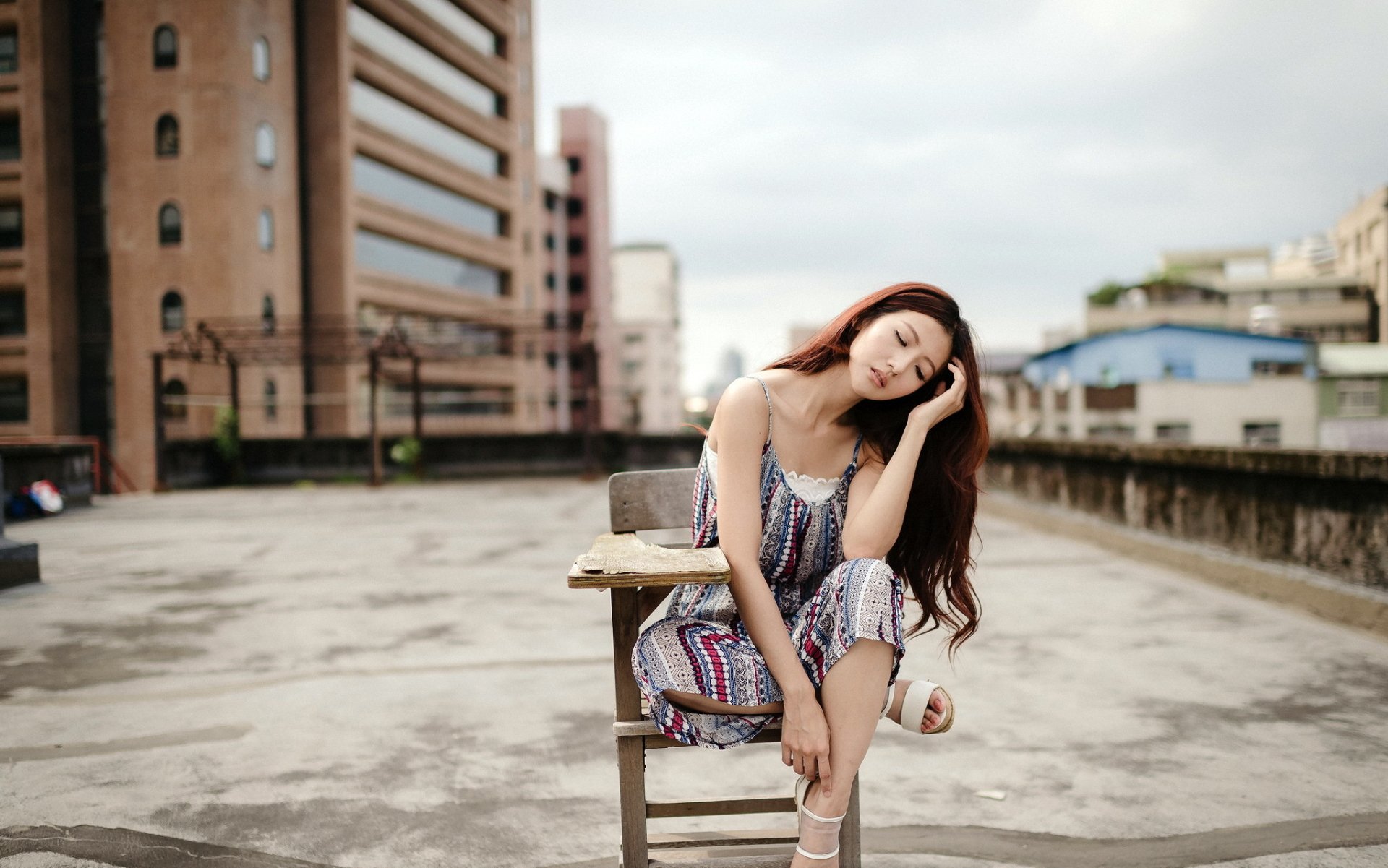 HD wallpaper featuring an Asian brunette woman sitting outdoors on a rooftop in a contemplative mood. The background includes urban buildings, creating an urban yet serene atmosphere.