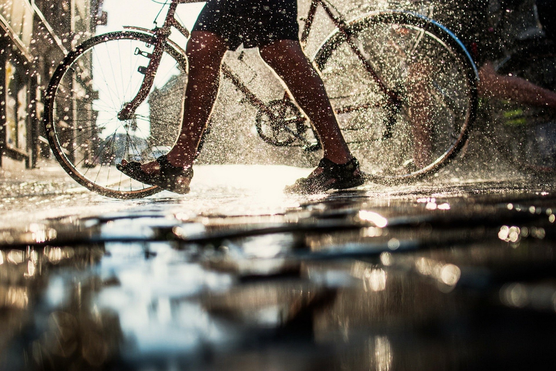 Artistic HD PC desktop wallpaper: sunlit legs and a bicycle splashing through rain-soaked puddles, water droplets sparkling around a walking human.