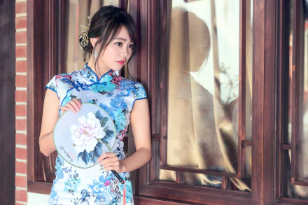 A brunette woman with brown eyes, wearing a blue floral kimono and lipstick, holds a decorative fan. She stands elegantly by a window, making for a beautiful HD desktop wallpaper and background.