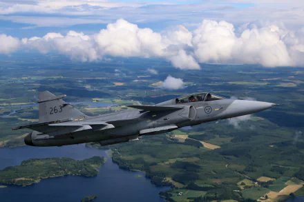 HD desktop wallpaper featuring a Saab JAS 39 Gripen jet fighter soaring above a scenic landscape with lakes and farmland under a partly cloudy sky.