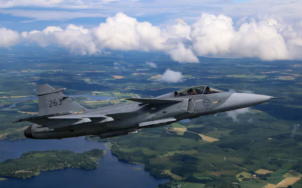 HD desktop wallpaper featuring a Saab JAS 39 Gripen jet fighter soaring above a scenic landscape with lakes and farmland under a partly cloudy sky.